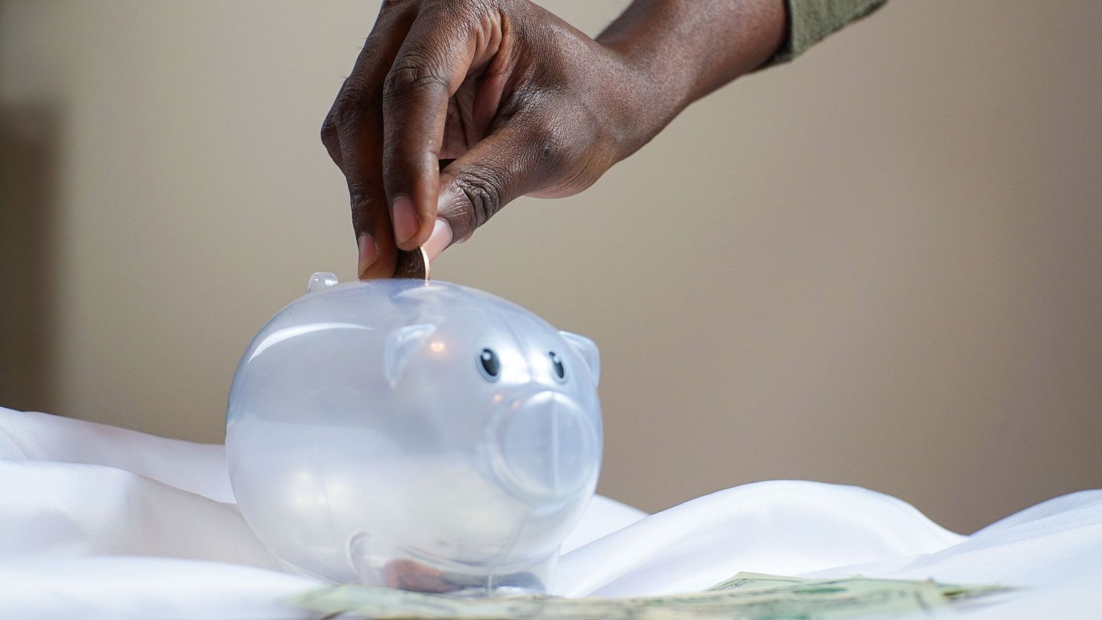 Hand dropping a coin into a clear piggy bank on a white cloth.